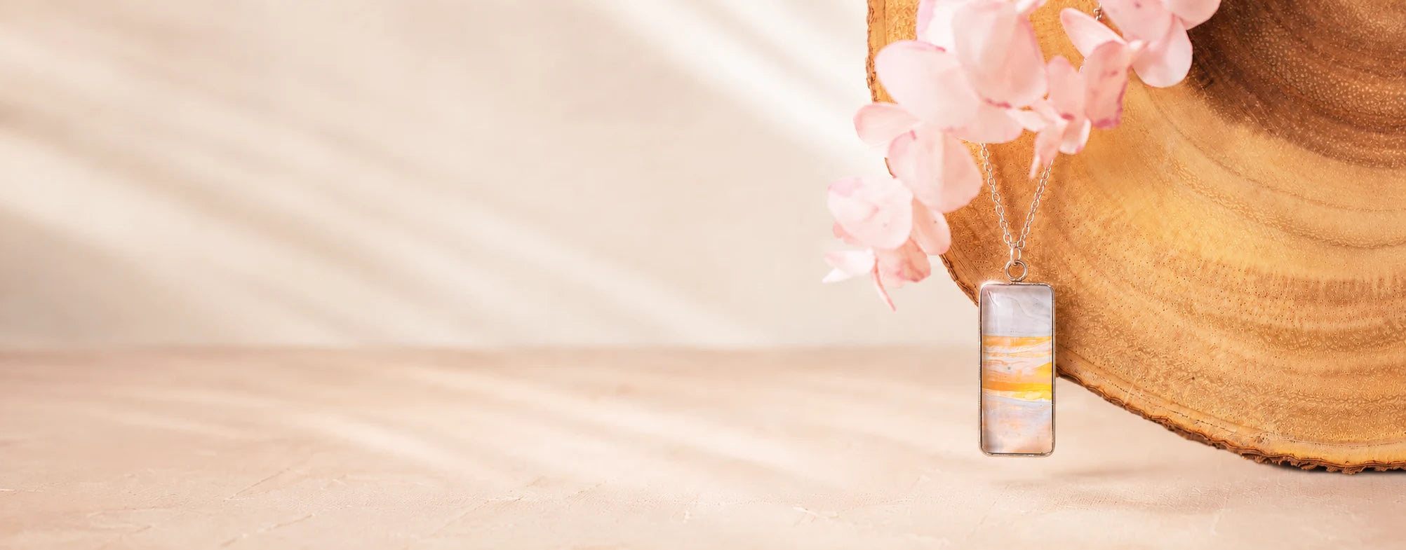 Dar Suad Stainless Steel rectangular marbled necklace in soft white, beige, and golden yellow displayed on a natural wood slice with delicate pink flowers against a light neutral background.